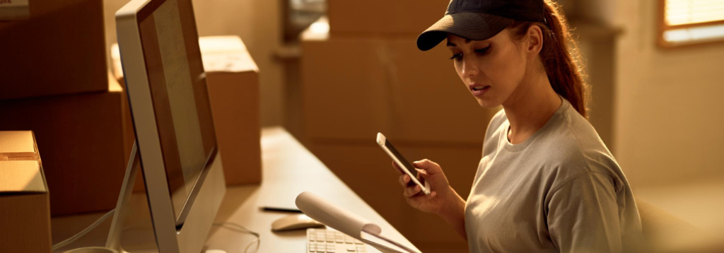 Mulher usando boné e camiseta em ambiente de estoque ou expedição, sentada em frente ao computador enquanto consulta o celular, com caixas de papelão ao redor.