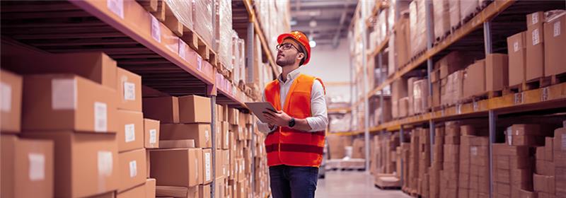 Profissional com colete e capacete no centro de distribuição, usando sistema de gestão para organizar estoque e pedidos.