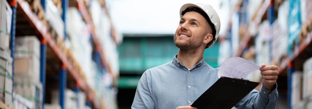 Profissional com capacete em corredor de armazém analisando planilha em prancheta, entre prateleiras com produtos, representando controle integrado de estoque e vendas.