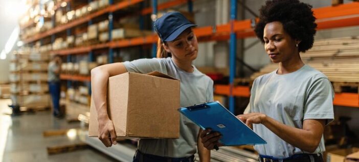 Duas mulheres trabalhando em um armazém. Uma delas segura uma caixa de papelão, enquanto a outra aponta para uma prancheta azul. Ambas usam camisetas cinzas e calças escuras. Ao fundo, há prateleiras altas cheias de caixas e materiais.