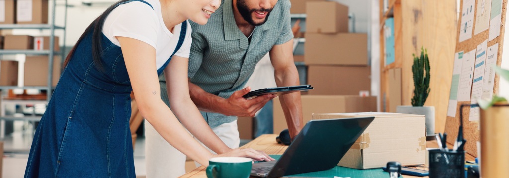 Profissionais em um armazém utilizando notebook e tablet para controlar produtos e pedidos, ilustrando o uso de software para gestão de estoque.